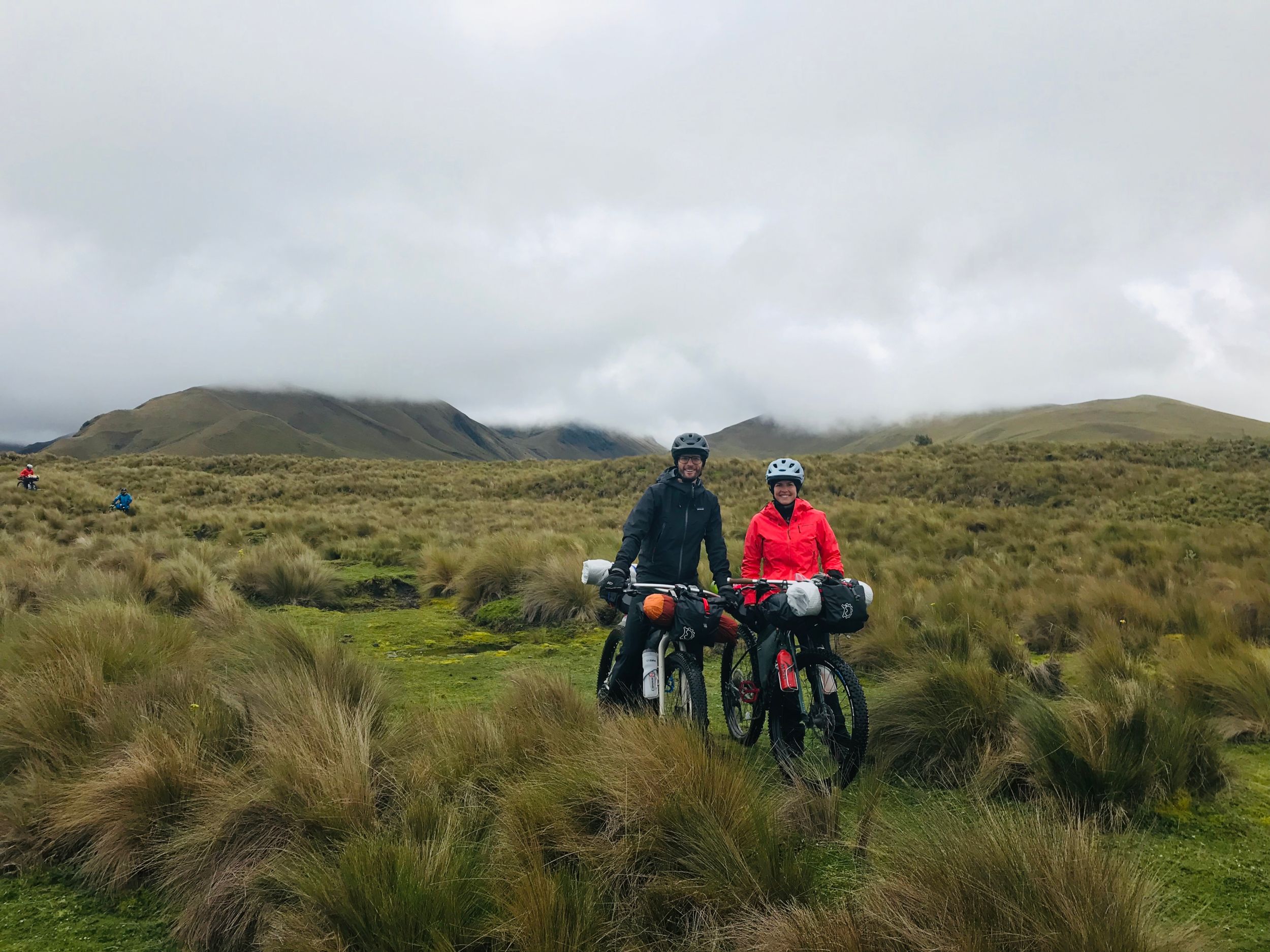 Two smiling people stand in a wet grassy field on a cloudy day
