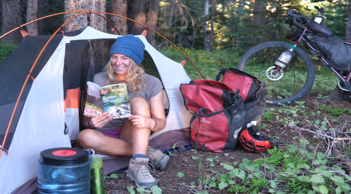 a young woman sits in front of a tent reading a book